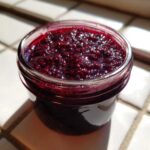 Close-up of a small glass jar filled with thick, dark purple elderberry syrup recipe concentrate, glistening in sunlight.
