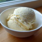Two scoops of creamy vanilla protein ice cream served in a small white bowl on a wooden surface.