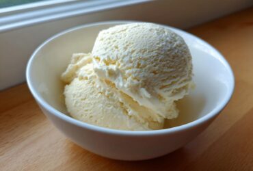 Two scoops of creamy vanilla protein ice cream served in a small white bowl on a wooden surface.