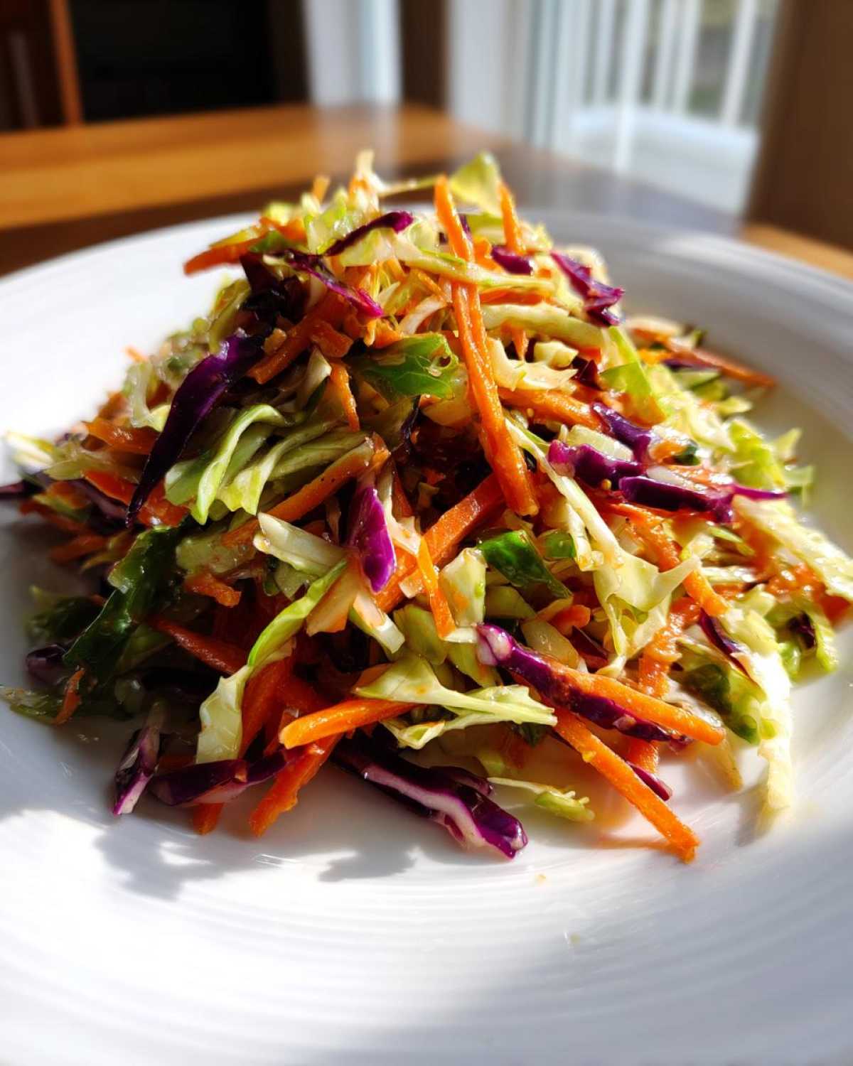 A vibrant mound of fresh cabbage salad featuring shredded green cabbage, purple cabbage, and bright orange carrots, served on a white plate.