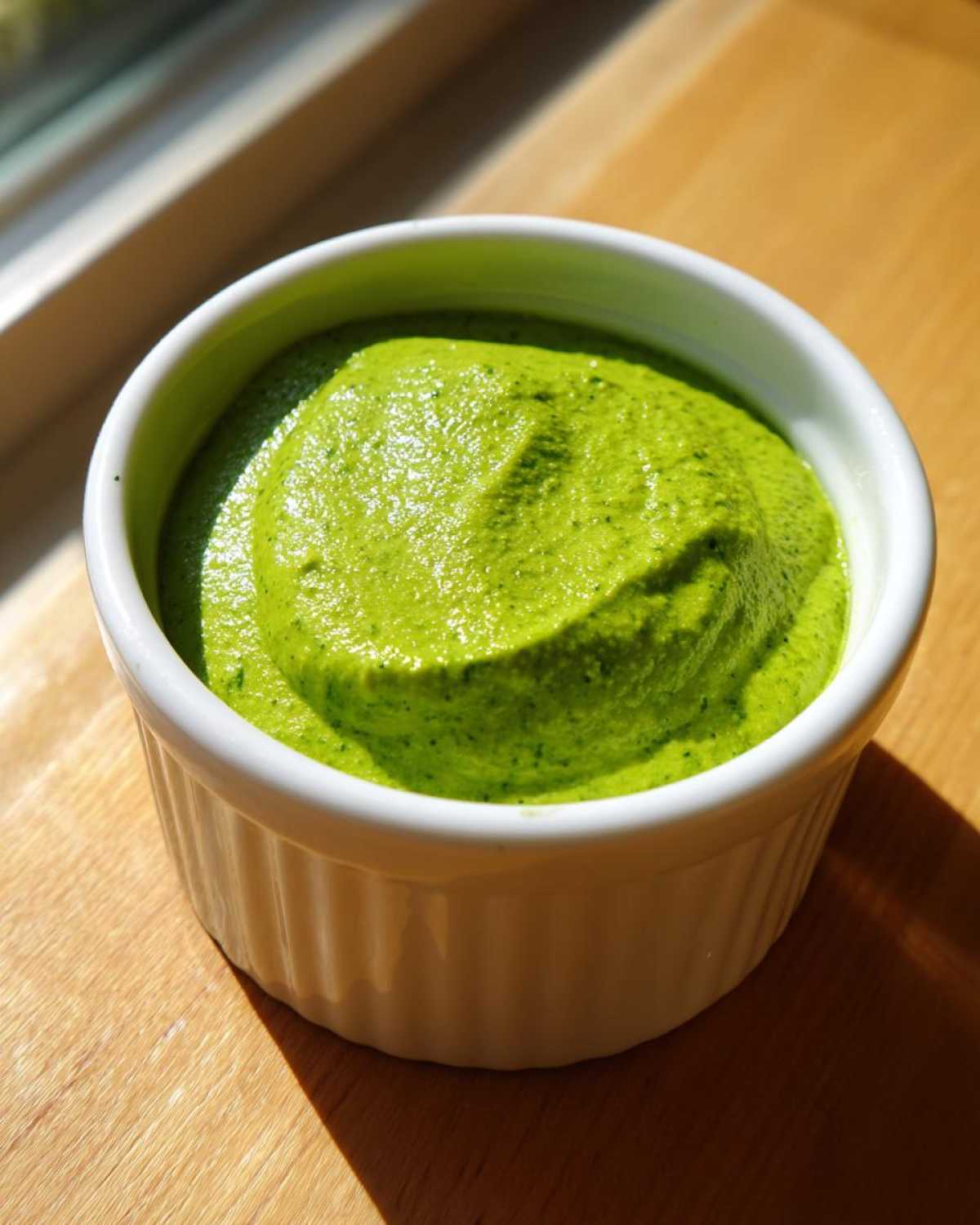 A close-up of vibrant green cilantro lime sauce served in a small white fluted ramekin on a wooden surface.
