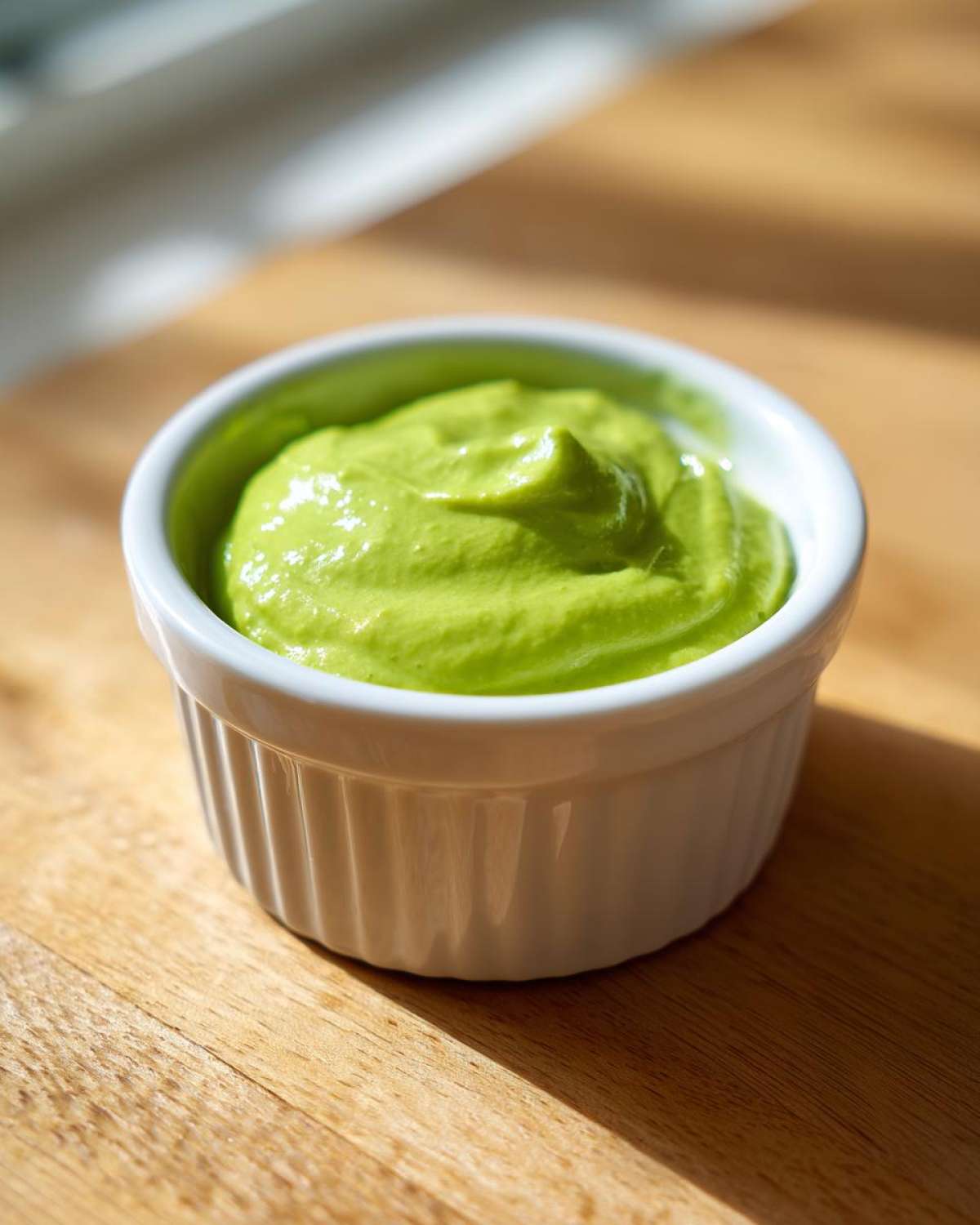 A close-up of bright green, creamy cilantro lime sauce served in a small white fluted ramekin on a wooden surface.