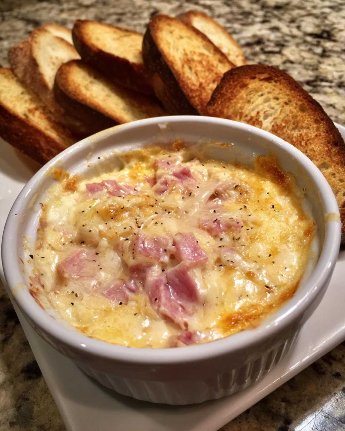 Close-up of a warm, cheesy reuben dip topped with diced ham, served with toasted bread slices.