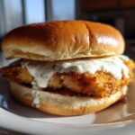 A close-up of an Air Fryer Crispy Fish Sandwich featuring a golden, breaded fillet generously topped with creamy tartar sauce on a brioche bun.