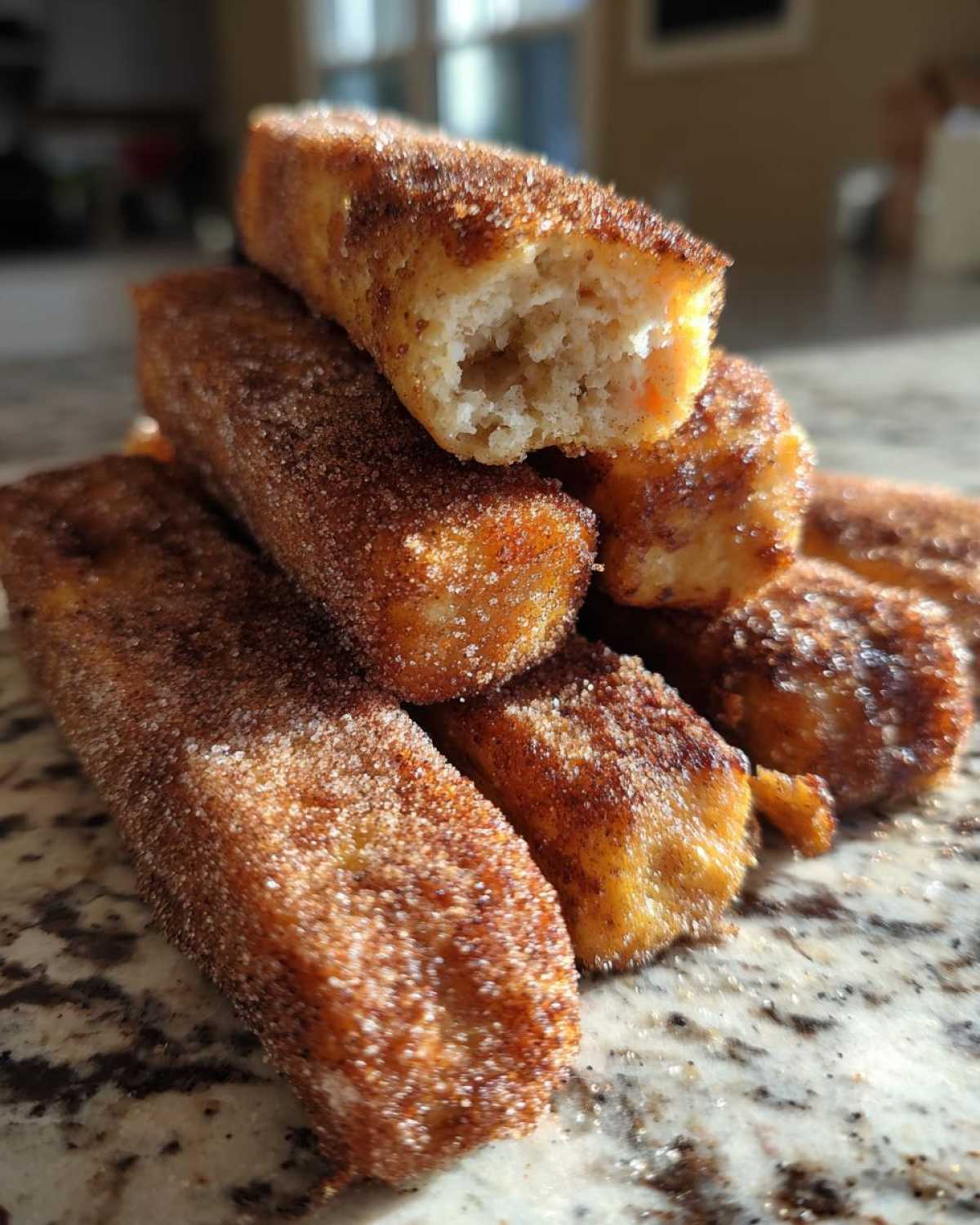 A stack of golden Air Fryer French Toast Sticks heavily coated in cinnamon sugar on a granite counter.