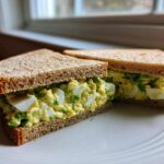 Two halves of an Avocado Egg Salad Sandwich made with whole wheat bread, showing creamy filling and chunks of egg white.