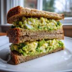A tall, stacked Avocado Egg Salad Sandwich made with whole wheat bread, showing chunky avocado and egg filling.