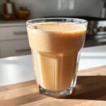 A tall glass filled with a thick, frothy Banana Peanut Butter Smoothie sitting on a wooden cutting board.