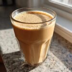 A tall glass filled with a creamy, light brown Banana Peanut Butter Smoothie sitting on a granite countertop near a window.