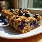 Close-up of a moist square of Blueberry Baked Oatmeal Squares, topped with fresh blueberries, served on a white plate.