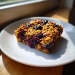 A single, thick Blueberry Baked Oatmeal Square with visible oats and jammy blueberries on a white plate.