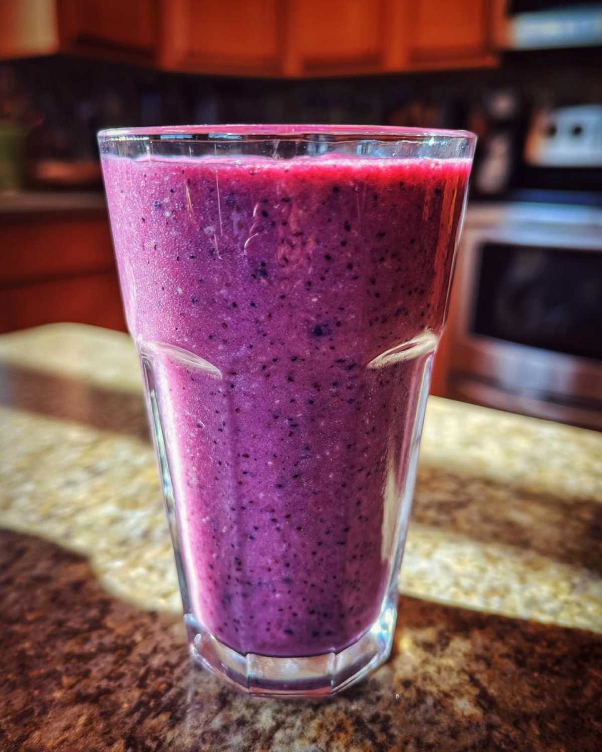A tall glass filled with a thick, vibrant purple Blueberry Lemon Chia Smoothie sitting on a granite countertop.
