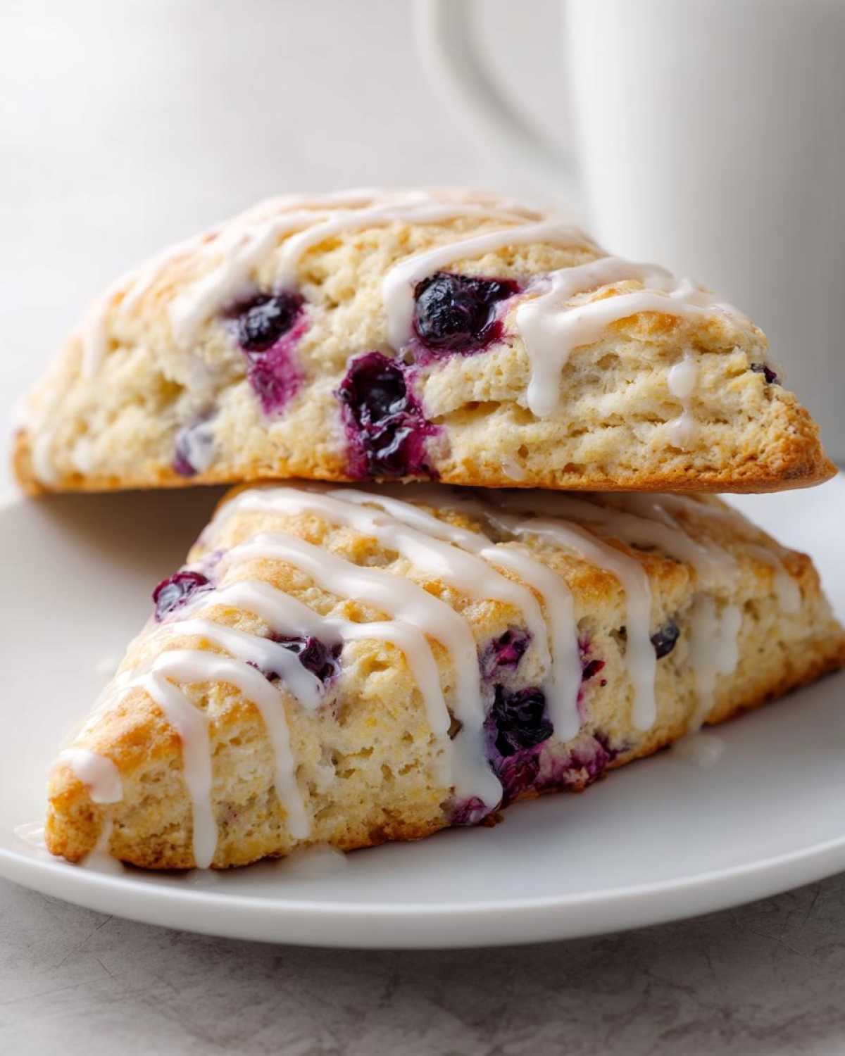 Two stacked Blueberry Lemon Scones drizzled with white lemon glaze, featuring visible blueberries.