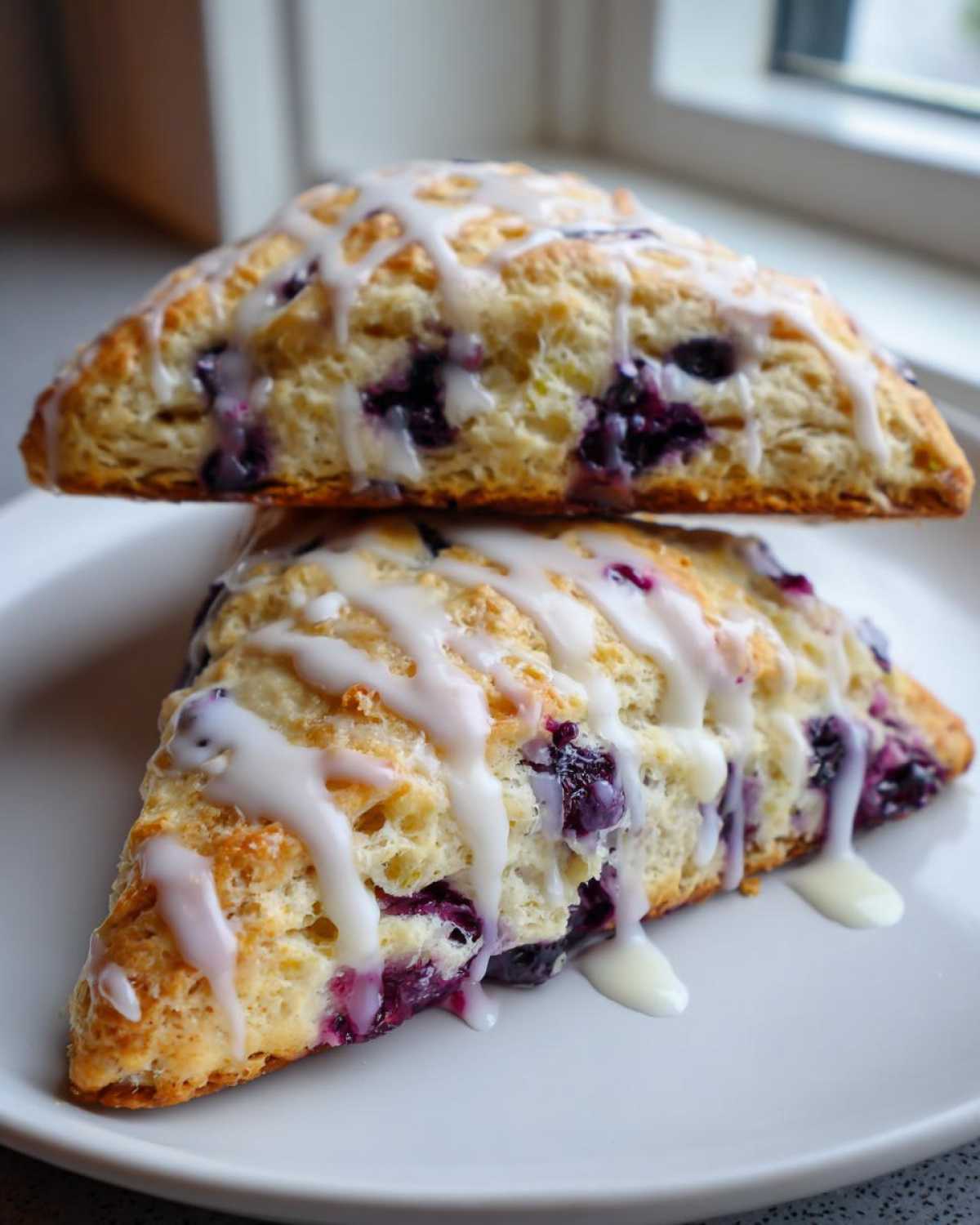 Two triangular Blueberry Lemon Scones stacked on a white plate, drizzled heavily with white lemon glaze.