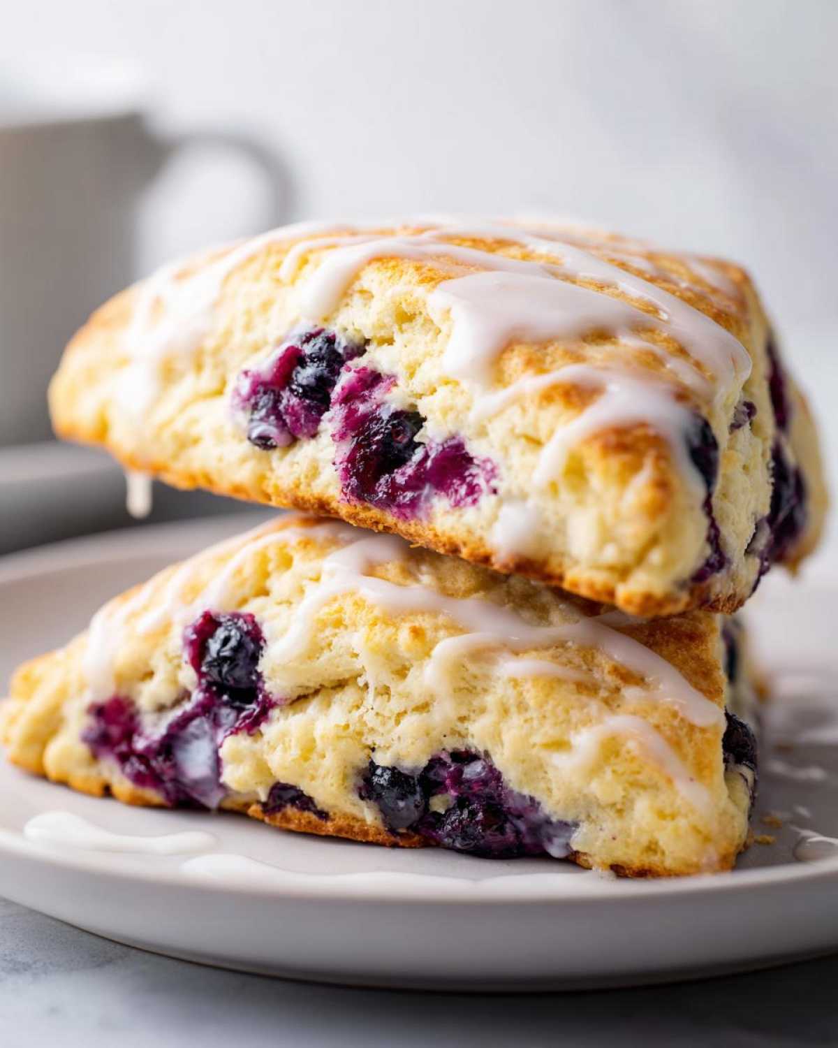 Two stacked, freshly baked Blueberry Lemon Scones topped with a sweet white glaze, showing juicy blueberries inside.