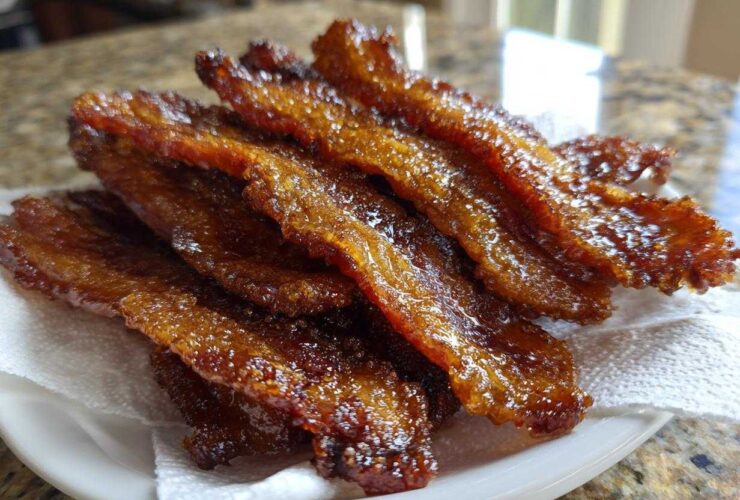 A stack of glistening, caramelized Brown Sugar Glazed Bacon resting on a white paper towel on a plate.