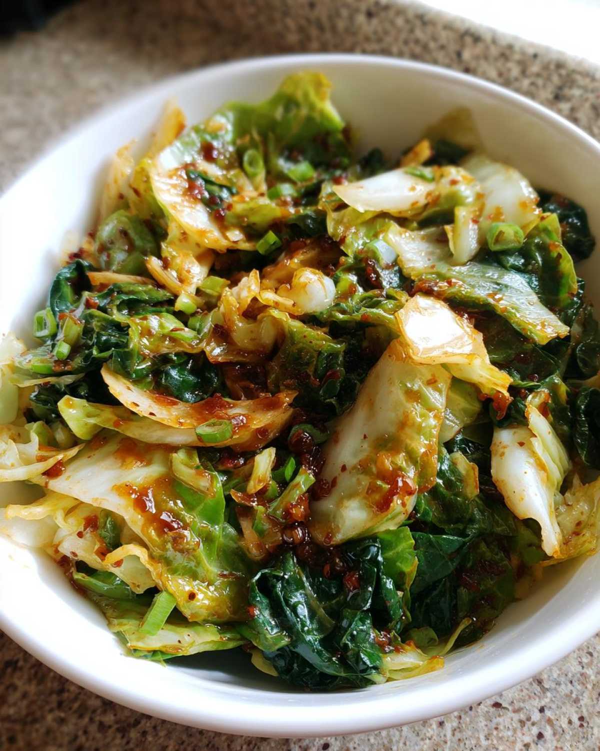 A close-up of a white bowl filled with Cabbage Stir Fry with Garlic Ginger Sauce, featuring glossy, wilted cabbage and green onions.