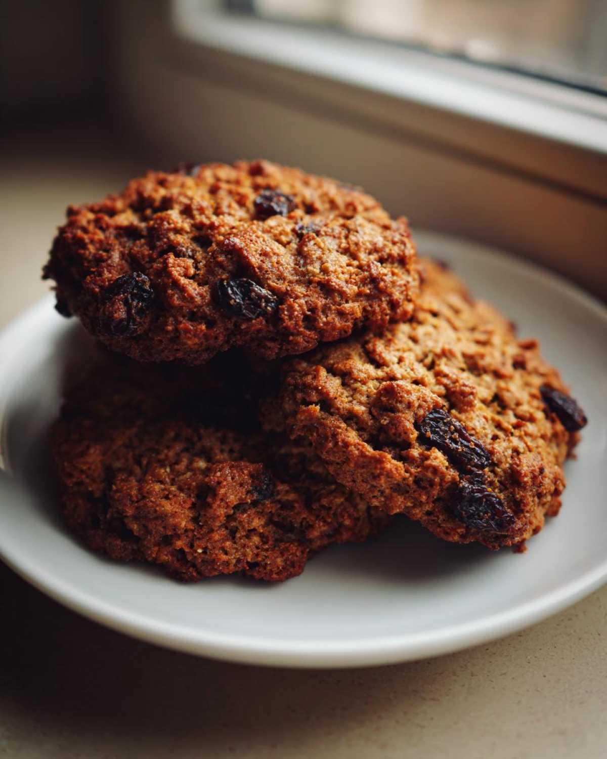 Close-up of three chewy Gluten-Free Oatmeal Raisin Cookies stacked on a white plate near a window.