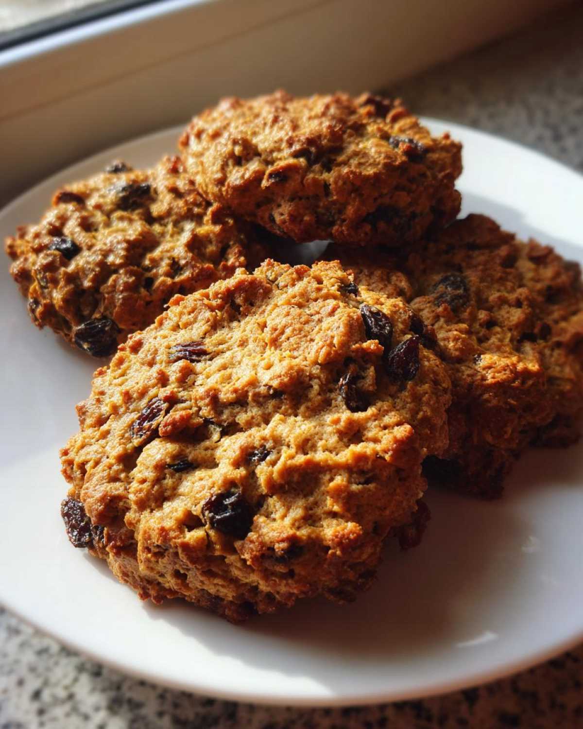 A stack of four golden-brown, chewy Gluten-Free Oatmeal Raisin Cookies piled on a white plate, illuminated by sunlight.