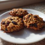 Three chewy Gluten-Free Oatmeal Raisin Cookies piled on a white plate, bathed in sunlight.