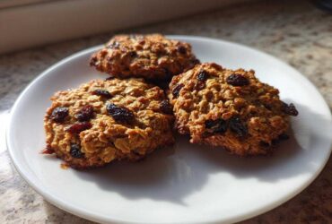 Three chewy Gluten-Free Oatmeal Raisin Cookies piled on a white plate, bathed in sunlight.