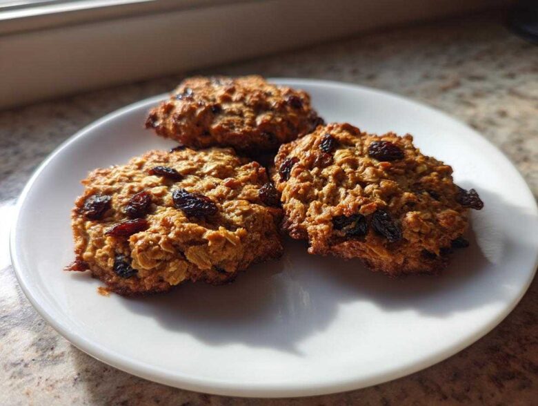 Three chewy Gluten-Free Oatmeal Raisin Cookies piled on a white plate, bathed in sunlight.