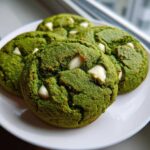 Four vibrant green, chewy matcha white chocolate cookies studded with white chocolate chips, resting on a white plate near a window.