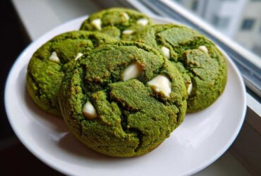 Four vibrant green, chewy matcha white chocolate cookies studded with white chocolate chips, resting on a white plate near a window.