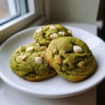 Three vibrant green Chewy Matcha White Chocolate Cookies studded with white chocolate chips, resting on a white plate near a window.