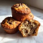 A stack of moist Chocolate Chip Banana Muffins, with one muffin broken in half showing the soft interior and chocolate chips.