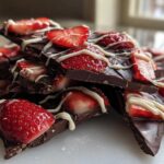 A stack of broken pieces of dark Chocolate Covered Strawberry Bark topped with fresh strawberry slices and white chocolate drizzle.