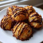 A close-up of golden brown Coconut Macaroons with Dark Chocolate Drizzle stacked on a white plate.