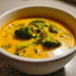 Close-up of a white bowl filled with thick, orange Copycat Broccoli Cheddar Soup topped with bright green broccoli florets.
