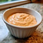 A small white ramekin filled with creamy, orange-tinted Copycat Raising Cane’s Sauce, showing visible specks of seasoning.