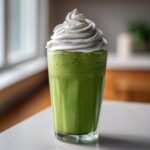 A tall glass filled with a vibrant green Copycat Shamrock Shake topped with a swirl of white mint whipped cream.