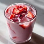 A refreshing glass of Copycat Starbucks Pink Drink, pink and creamy, topped with ice, sliced strawberries, and dried rose petals.