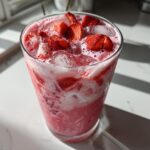 Close-up of a glass filled with a pink, creamy beverage, ice, and fresh strawberry pieces, resembling a Copycat Starbucks Pink Drink.