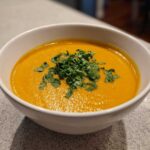 A close-up of a white bowl filled with vibrant orange Creamy Coconut Lentil Soup, topped generously with fresh chopped cilantro.