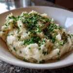 A close-up of a bowl filled with fluffy Creamy Garlic Mashed Cauliflower, topped generously with chopped green chives and pepper.