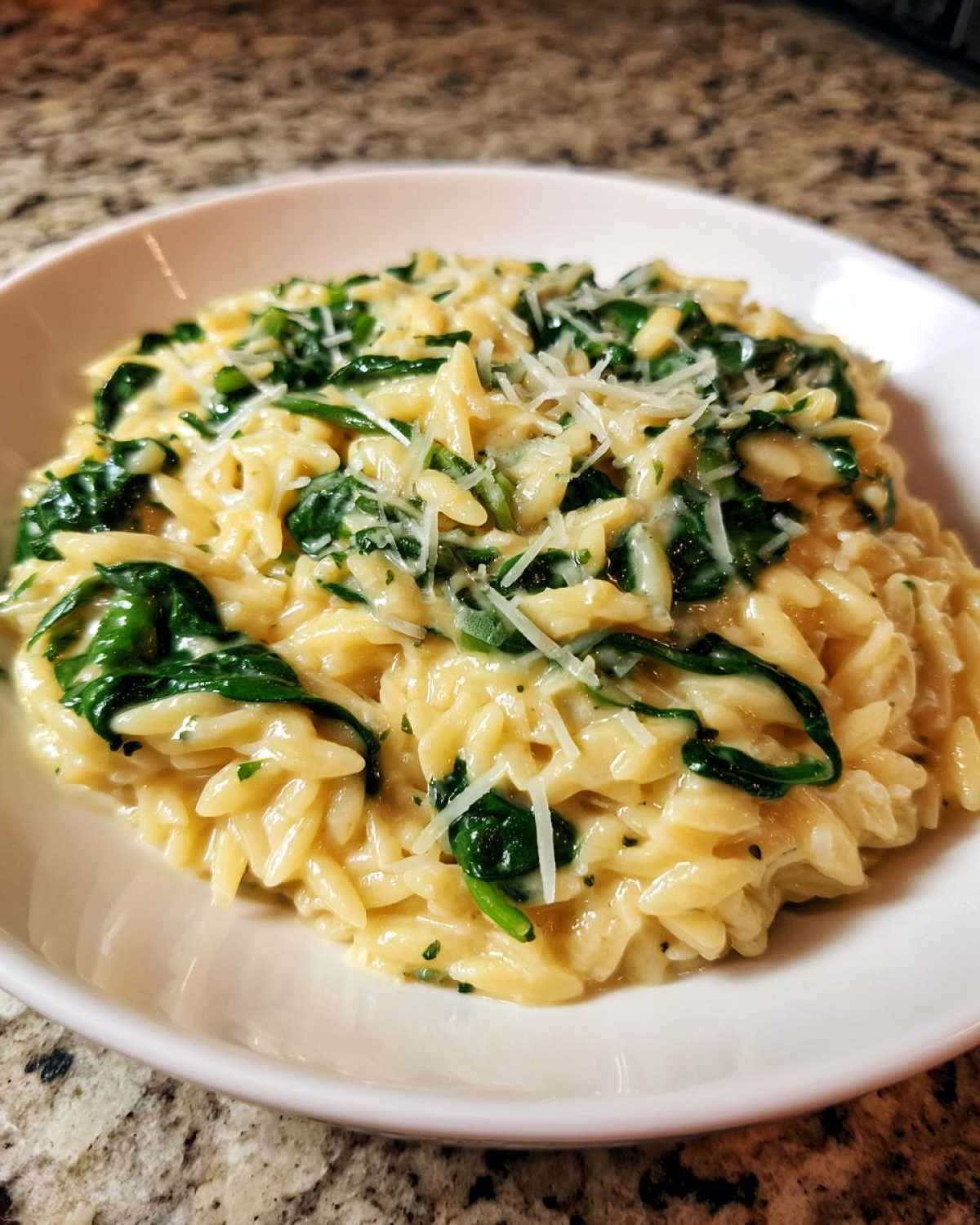 A close-up of a white bowl filled with Creamy Garlic Parmesan Orzo with Spinach, topped with shredded cheese.
