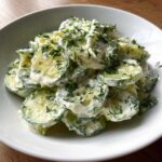 A white bowl filled with sliced cucumbers coated in a creamy dressing and topped generously with fresh dill, showcasing the Creamy German Cucumber Salad.
