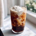 A tall glass showing a swirling, layered Iced Mocha with ice cubes and topped generously with whipped cream.