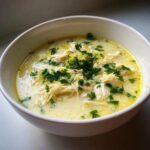 A close-up of a white bowl filled with Creamy Lemon Chicken Orzo Soup, topped with fresh parsley and lemon zest.
