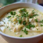 Close-up of a white bowl filled with Creamy Lemon Chicken Orzo Soup, topped with shredded chicken and fresh parsley.