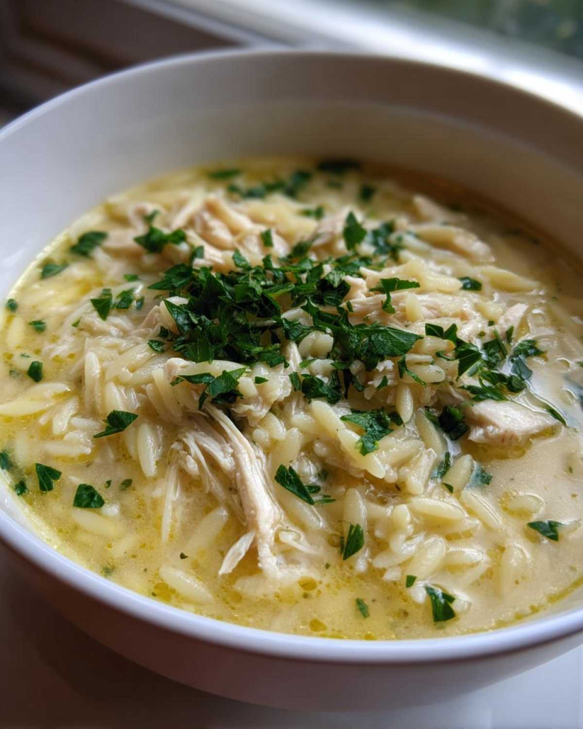 A close-up view of a bowl of Creamy Lemon Chicken Orzo Soup garnished with fresh parsley.