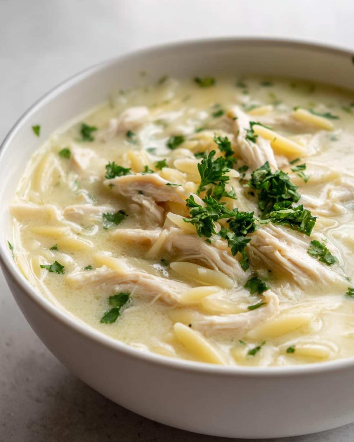 A close-up shot of a bowl of rich Creamy Lemon Chicken Orzo Soup, topped with shredded chicken and fresh parsley.