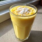 A tall glass of bright yellow Pineapple Coconut Smoothie topped with shredded coconut flakes.