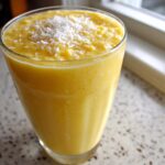 A tall glass filled with a thick, bright yellow Pineapple Coconut Smoothie, topped with shredded coconut.