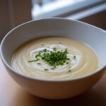 A close-up of a white bowl filled with smooth, creamy potato leek soup, garnished with fresh chives and a swirl of cream.