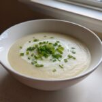 A white bowl filled with smooth, creamy potato leek soup, garnished with a swirl of cream and fresh chopped chives.